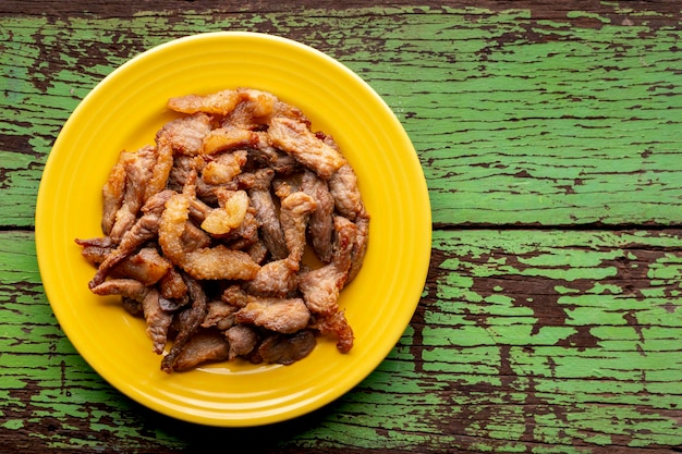 Saborosa carne de porco frita em placa de cerâmica amarela sobre fundo de textura de madeira natural verde velho rústico com espaço de cópia para texto vista superior comida de rua plana leiga