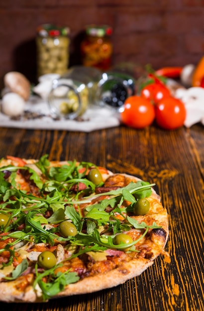 Saborosa pizza com rúcula e azeitonas na mesa de madeira, tomates, cogumelos e outros vegetais no fundo