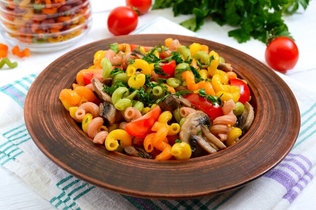 Saborosa salada quente. Macarrão colorido com cogumelos e tomates frescos em uma tigela sobre uma superfície de madeira branca. Macarrão colorido.