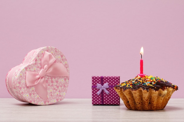 Saboroso bolinho de aniversário com esmalte de chocolate e caramelo, decorado com velas festivas acesas e caixas de presente em fundo violeta. Conceito mínimo do dia de celebração.