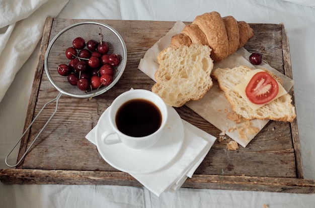 Saboroso café da manhã com croissant fresco, café, cerejas em uma bandeja de madeira. Croissant saudável com tomate e queijo. Café expresso na bandeja de café da manhã