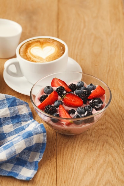 Saboroso café da manhã com iogurte grego leve, morangos frescos, framboesas, mirtilos, amoras e xícara de café espumoso quente. Refeições matinais com baixo teor de gordura e início de dia saudável. Conceito de desintoxicação e dieta