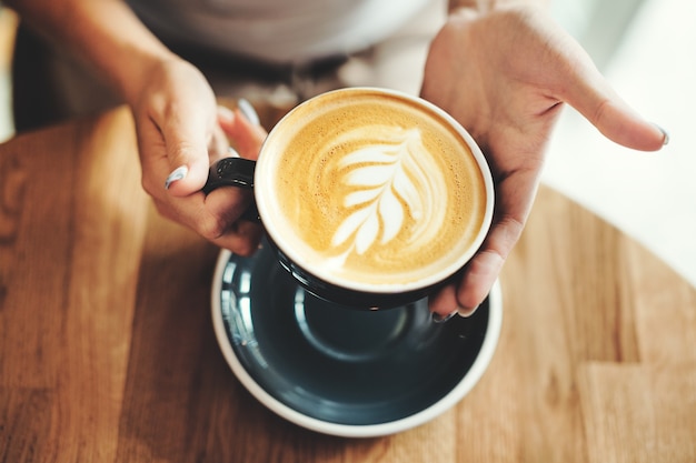 Saboroso cappuccino fresco na xícara na mesa de madeira. mulher irreconhecível segurando a taça nas mãos.