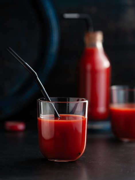 Saboroso suco de tomate derramado em copos com um canudo de coquetel, uma garrafa de suco no escuro