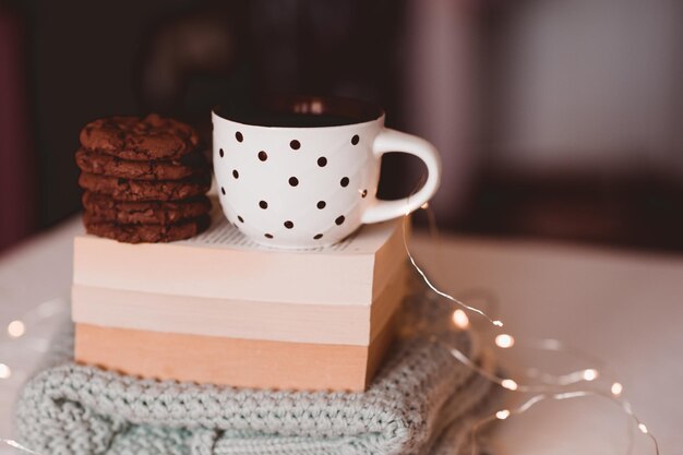 Saborosos biscoitos de chocolate com chá quente fresco em livros abertos e closeup de suéter de malha sobre as luzes de Natal na sala. Bom Dia. Café da manhã. Hora da Escola.