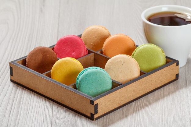 Saborosos bolos de macaroons de cores diferentes em uma caixa de madeira marrom e uma xícara de café em placas cinza