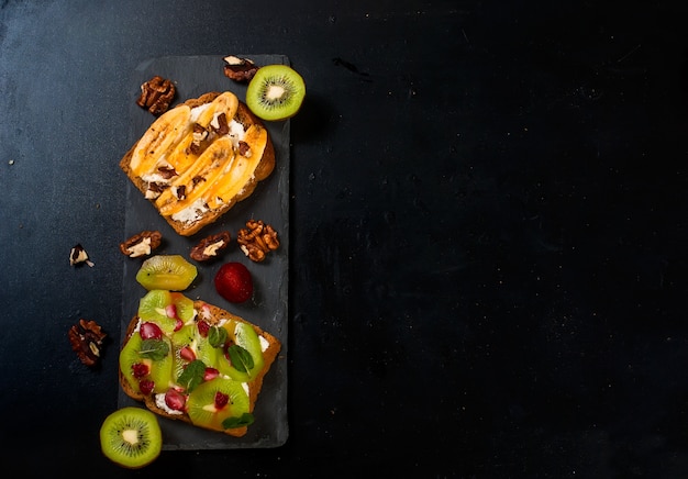 Saborosos doces sanduíches com bananas, nozes e chocolate, kiwi, morangos e hortelã em fundo preto