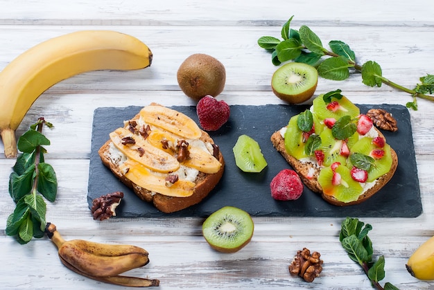 Saborosos doces sanduíches com bananas, nozes e chocolate, kiwi, morangos e hortelã na mesa de madeira