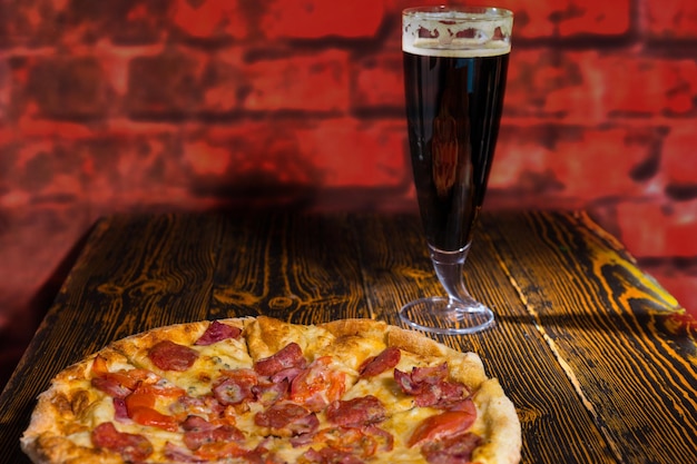 Sabrosa pizza de pepperoni con variedad de ingredientes y queso en la mesa de madera cerca de un vaso de cerveza oscura