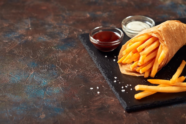 Sabrosas papas fritas con salsas en la tabla de cortar de pizarra, sobre fondo oscuro