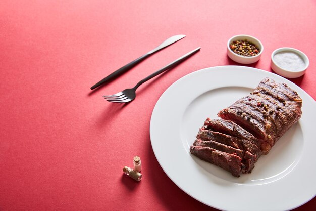 Sabroso bistec a la parrilla servido en un plato con cubiertos y sal y pimienta en tazones sobre fondo rojo