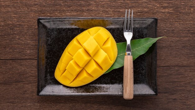 Sabroso mango maduro amarillo fresco con tenedor y hoja de mango en plato de cerámica rectangular negro