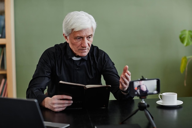 Sacerdote mayor de pelo blanco sosteniendo la biblia y grabando video