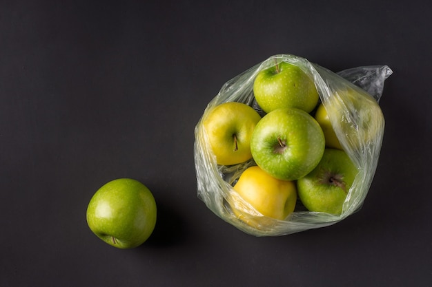 Saco de celofane amassado de plástico transparente com um grupo de maçãs verdes e amarelas maduras em um fundo preto