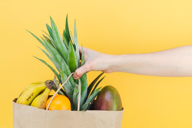 Saco de compra de papel cheio de frutas tropicais com a mão de uma mulher em fundo amarelo. Copie o espaço.