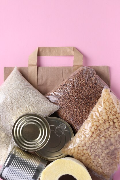 Saco de papel com suprimentos alimentares estoque de alimentos de crise para o período de isolamento de quarentena na superfície rosa. arroz, trigo sarraceno, macarrão, comida enlatada, papel higiênico. entrega de alimentos, doação, formato vertical, vista superior