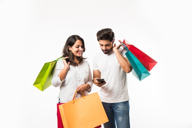 Sacos de compras do jovem casal indiano e telefone inteligente ou celular, clicando em selfie ou localizando loja, isolado sobre fundo branco. foco seletivo