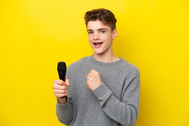 Sänger Teenager-Mann, der ein Mikrofon aufnimmt, das auf gelbem Hintergrund isoliert ist und einen Sieg feiert