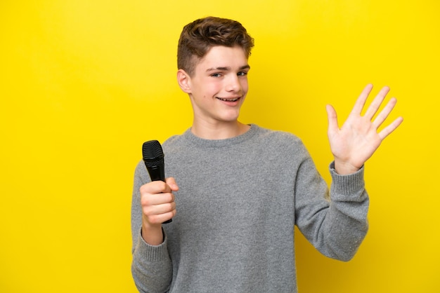 Sänger Teenager-Mann, der ein Mikrofon aufnimmt, das auf gelbem Hintergrund isoliert ist und mit der Hand mit glücklichem Ausdruck salutiert
