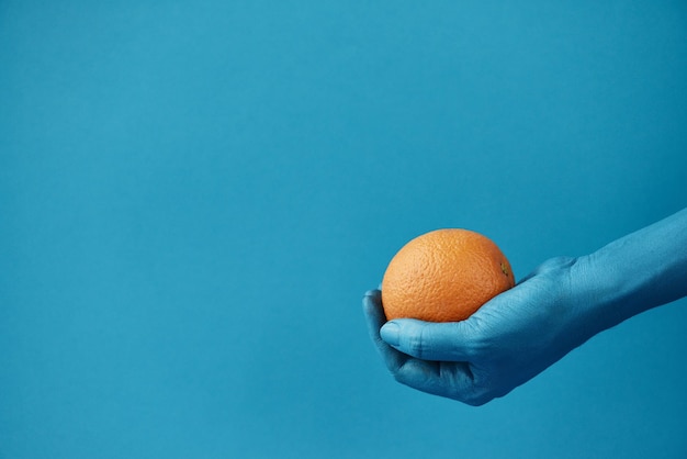 Saftige frische Orange, die in männlicher Hand liegt, die in Blau bemalt ist und sich vor türkisfarbenem Hintergrund abhebt