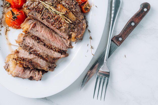 Saftige, gut gebratene Ribeye-Steaks auf einer weißen Platte auf einer weißen Marmoroberfläche mit Himbeere und gebratenen Kirschtomaten und Paprikasteaks sind in Scheiben geschnitten und servierfertig