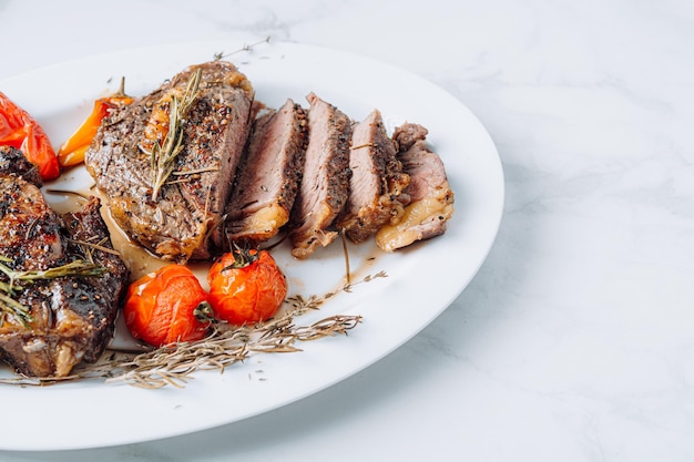 Saftige, gut gebratene Ribeye-Steaks auf einer weißen Platte auf einer weißen Marmoroberfläche mit Himbeere und gebratenen Kirschtomaten und Paprikasteaks sind in Scheiben geschnitten und servierfertig