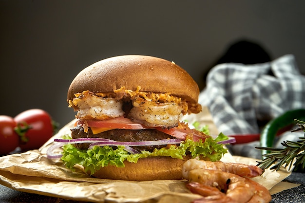 Saftiger Burger mit Rindfleisch, Garnelen, Tomaten, Käse und knusprigen Zwiebeln auf einem dunklen Tisch. Nahaufnahme, selektiver Fokus