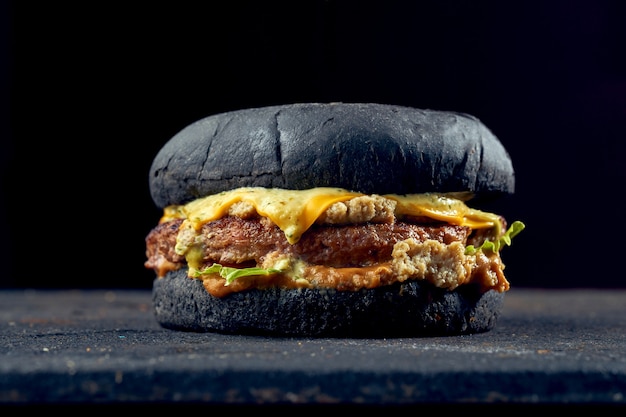 Saftiger Burger mit schwarzen Brötchen mit Rindfleisch, Käse, Gurke und Tomatensauce auf dunklem Hintergrund.