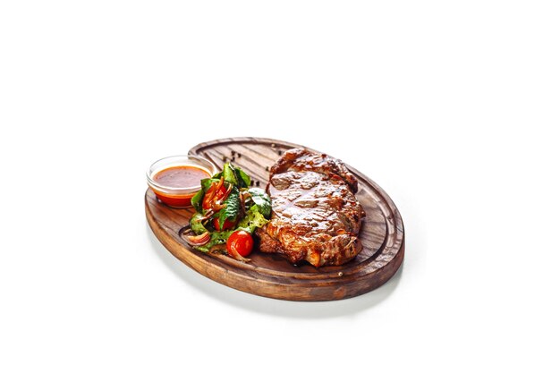 Saftiges appetitliches gegrilltes Steak, serviert mit einem Salat aus Tomaten und grünen Blättern und mit rotem Saucenessen, serviert auf einem Holzbrett, isoliert