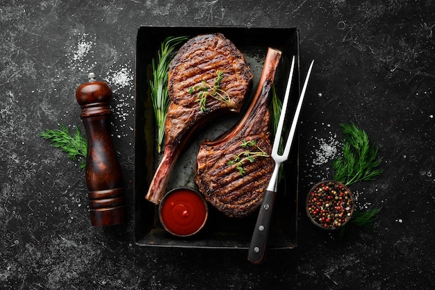 Saftiges Steak am Knochen gegrillt mit Gewürzen und Kräutern auf schwarzem Steinhintergrund Draufsicht Freier Kopierplatz