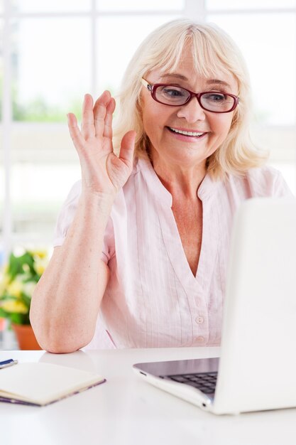 Sagen hallo zu ihrem Nächsten. Fröhliche ältere Frau winkt mit der Hand beim Betrachten des Laptops