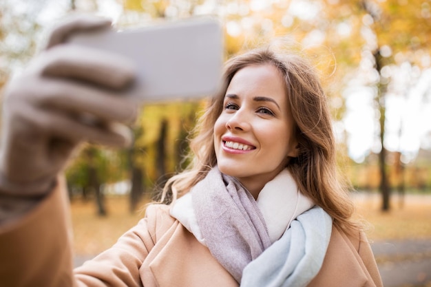 Saison-, Technologie- und Menschenkonzept - schöne junge glückliche Frau, die im Herbstpark ein Selfie mit dem Smartphone macht