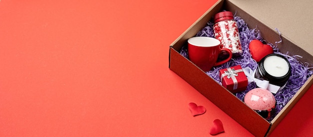 Saisonale Geschenkbox für den Valentinstag mit kerzenroter Tasse und herzförmigen Süßigkeiten auf rotem Hintergrundbanner