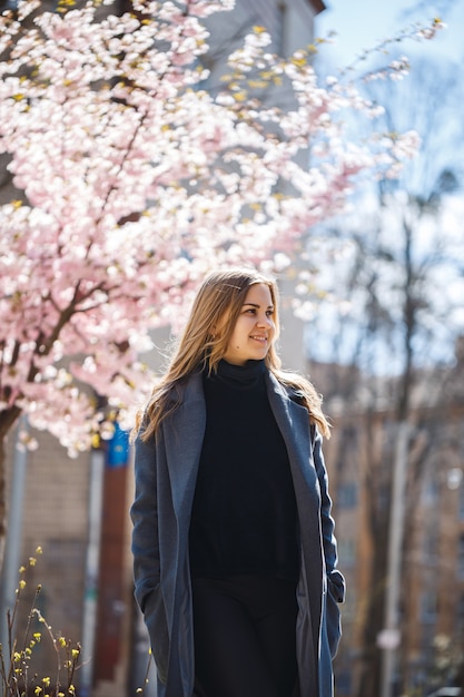 Sakura se ramifica com flores em uma árvore nas ruas da cidade. Menina mulher feliz em uma paleta cinza caminha ao longo de um beco com sakura florescendo. Linda garota chique ao ar livre. Árvore de Sakura florescendo.