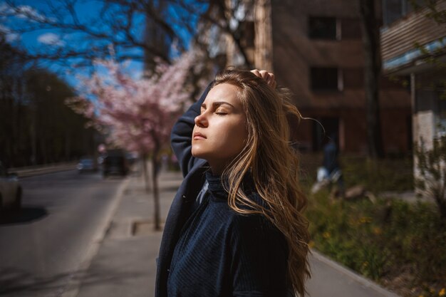 Sakura verzweigt sich mit Blumen an einem Baum auf den Straßen der Stadt. Glückliches Frauenmädchen, das sich mit blühender Kirschblüte auf der Straße dreht. Wunderschönes modisches Mädchen im Freien. Sakura-Baum blüht.