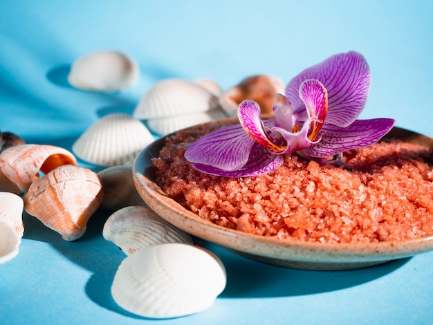 Sal de baño naranja en un platillo con conchas y flores sobre un fondo azul con una sombra de una planta tropical. Copyspace Spa, relajado, verano