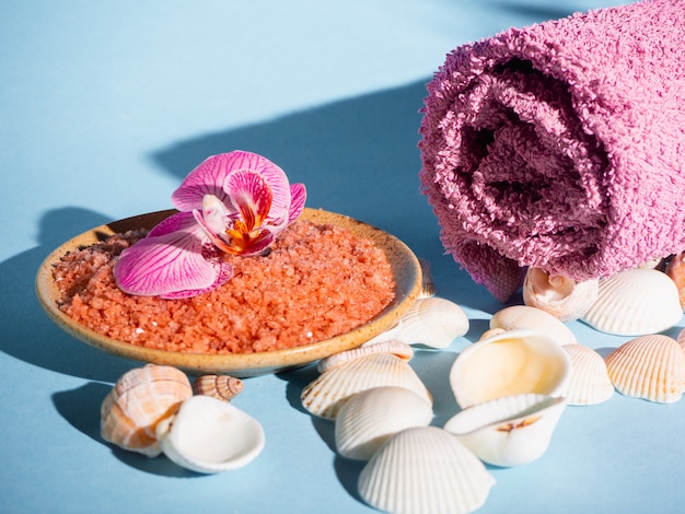 Foto sal de banho alaranjado em um pires com conchas, toalha e flor em um fundo azul com uma sombra de uma planta tropical. copyspace. spa, relaxado, verão