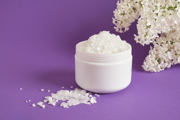 Sal do mar branco em uma jarra de plástico e flores lilás brancas em fundo lilás conceito cosmético para cuidados com o corpo natural