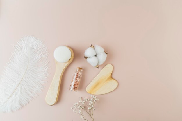 Una sal marina rosa en una pequeña botella de vidrio, un cepillo facial y un masajeador Guasha de madera sobre un fondo rosa