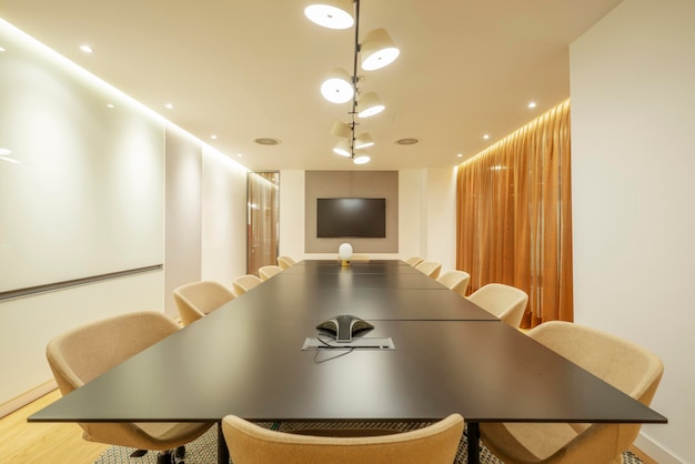 Sala de juntas con mesa negra larga de una oficina profesional con TV de pantalla plana en la pared y sillas giratorias negras con tapicería clara y cortinas marrones