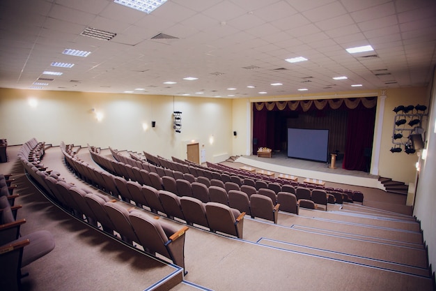 Sala de reuniones de conferencia con luces LED de techo, sillas de fila marrón, con escenario y pantalla vacía para reuniones de negocios