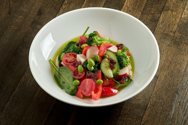 Salada apetitosa de vegetais com pepino, tomate, rabanete, feijão e brócolis com manteiga verde em uma tigela branca na mesa de madeira