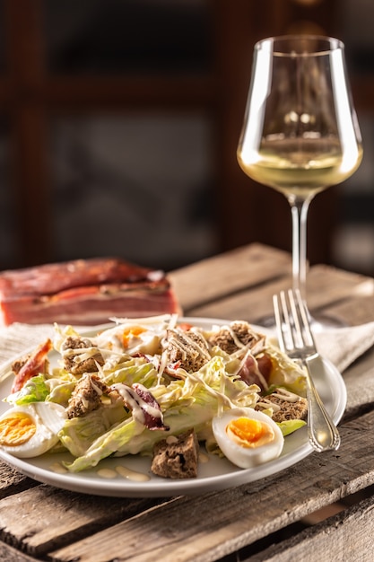 Salada caesar com ovos cozidos, bacon e croutons servidos em um prato ao lado de uma taça de vinho branco.
