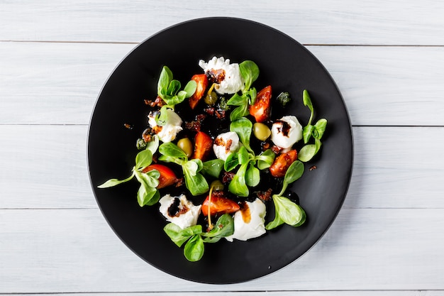 Salada caprese italiana fresca com mussarela e tomate no prato escuro na mesa de madeira branca.