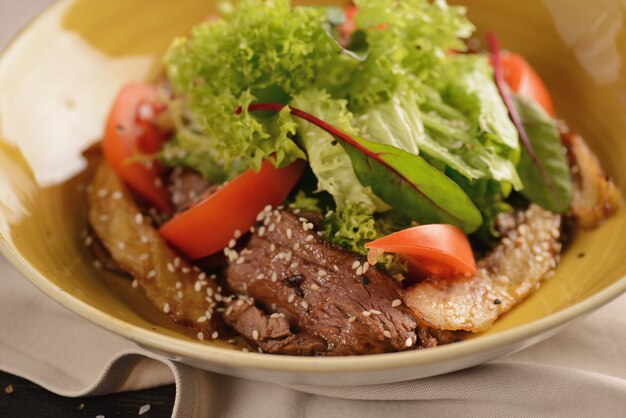 Salada com carne, tomate, sementes de gergelim e alface. Em um prato amarelo em uma mesa de madeira