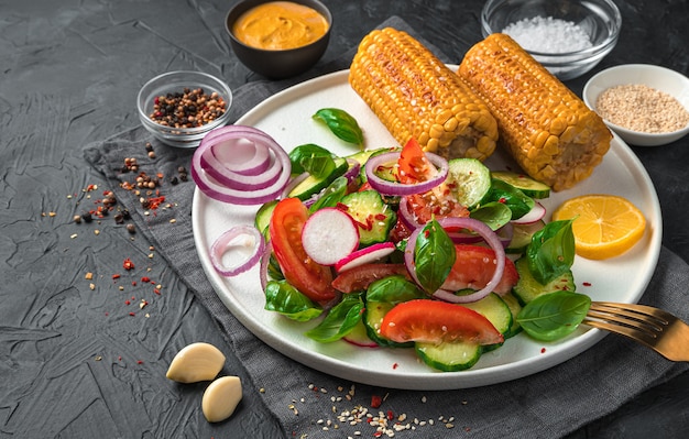 Salada com legumes frescos, tomates, pepinos, rabanetes e milho frito em um prato branco sobre uma parede escura