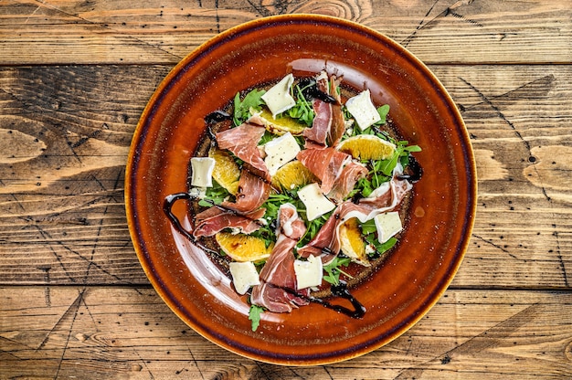 Salada com presunto de parma prosciutto, queijo parmesão, rúcula e tangerina no prato. Mesa de madeira. Vista do topo.