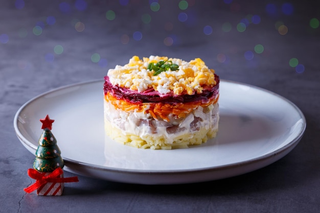 Salada de arenque sob um casaco de pele. Salada russa tradicional em várias camadas de arenque, beterraba, batata, cenoura e ovos. Close-up, fundo cinza, árvore de Natal em miniatura, bokeh.