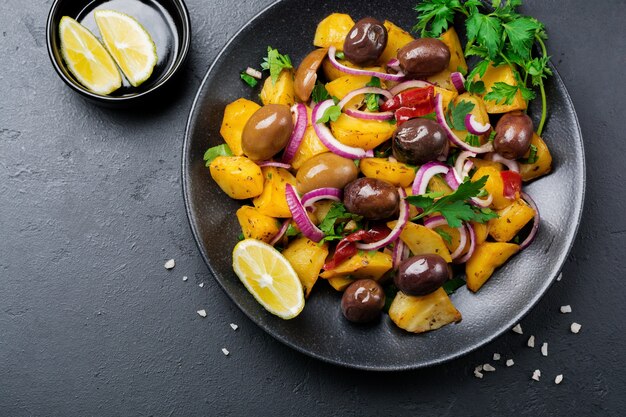 Salada de batata quente com azeitonas, pimenta, salsa e cebola roxa na placa de cerâmica preta sobre fundo escuro de concreto