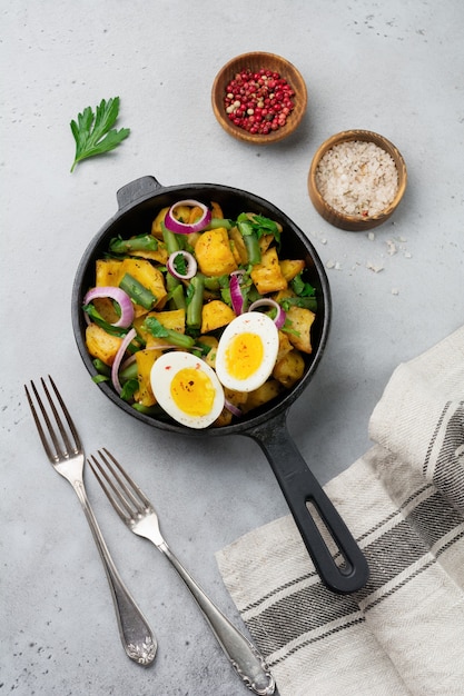 Salada de batata quente com feijão verde, pimenta, salsa, ovos e cebola roxa na frigideira na mesa de concreto cinza. Foco seletivo. Vista do topo. Copie o espaço.